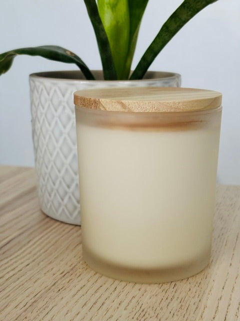 Frosted glass candle with wooden lid on a wooden surface, with a plant in the background.